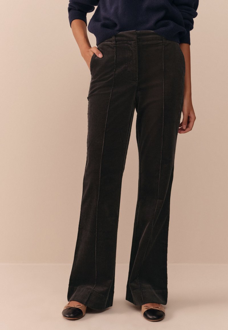 Pantaloni in velluto a coste scuro dal taglio dritto, con tasche frontali e cuciture verticali a contrasto. Indossati con ballerine nere.