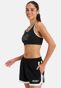Sujeción deportiva negra con acentos blancos y el logo de "ROXY". Combinada con pantalones cortos negros que tienen rayas blancas y detalles de bolsillo.