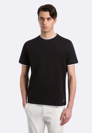 Giovane uomo con capelli scuri che indossa una maglietta nera a maniche corte con rifinitura bianca e pantaloni chiari, in piedi davanti a uno sfondo semplice.