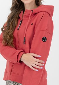 Felpa rossa con zip in tessuto morbido, con tasca frontale, cappuccio con cordino regolabile, patch con logo nero sulla manica e polsini elastici.