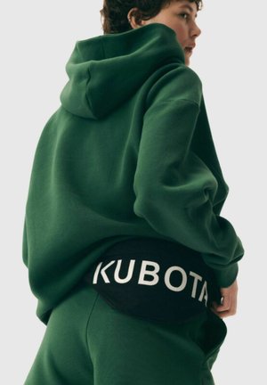 Zielona bluza z kapturem o luźnym kroju, zestawiona z czarnym pasem w talii z napisem "KUBOTA" dużymi białymi literami. Miękka tekstura materiału.