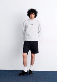 Light gray sweatshirt with embroidered text, paired with black shorts. Model wears black shoes and white socks, standing on blue carpet.