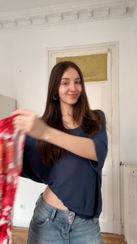 Νεαρή γυναίκα με μακριά καστανά μαλλιά που χαμογελά, κρατώντας ένα κόκκινο ύφασμα με μοτίβο, στέκεται σε ένα λευκό δωμάτιο με μια κλειστή πόρτα πίσω της.