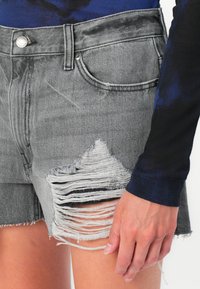 Shorts en denim gris avec un ourlet effiloché et des patchs abîmés, dotés d'une fermeture à bouton et d'un design à cinq poches. Texture lisse et coupe décontractée.