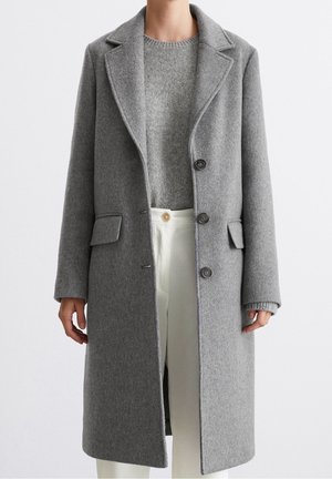 Frau trägt einen grauen Wollmantel mittlerer Länge über einem grauen Strickpullover und weißer, hoch taillierter Cordhose, steht vor einem schlichten Hintergrund.