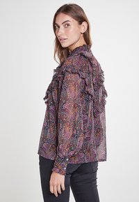 Blusa de estampado oscuro con volantes en el escote y la parte frontal, con un diseño de paisley en púrpura, rojo y amarillo sobre un fondo negro.