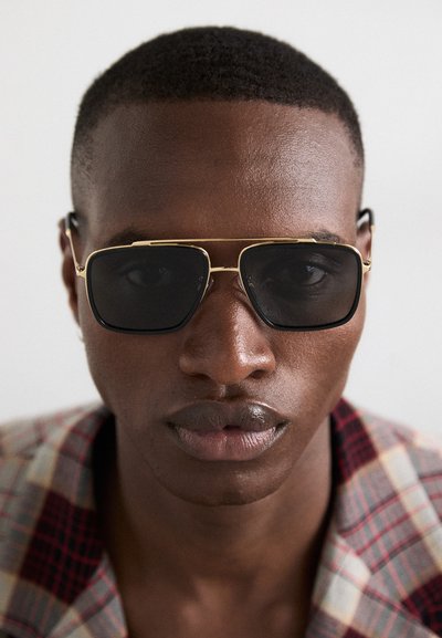 Hombre con gafas de sol oscuras de forma rectangular y chaqueta de cuadros, mirando hacia adelante sobre un fondo claro y liso.