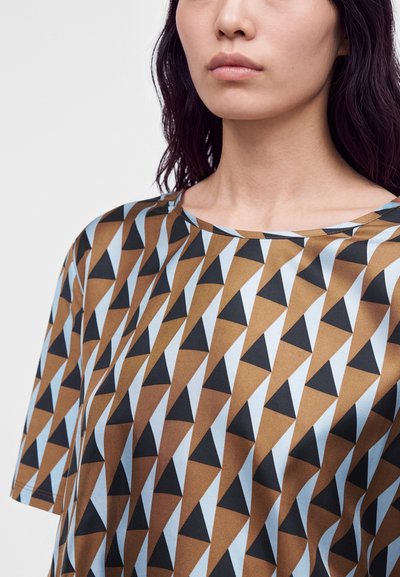 Femme portant une chemise à manches courtes avec un motif géométrique de triangles marron, noir et bleu clair sur fond blanc.