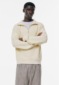 Cremefarbener, gerippter Reißverschluss-Pullover mit hohem Kragen und langen Ärmeln. Kombiniert mit hellgrauen gestreiften Hosen. Minimalistisches Design und weicher Stoff.