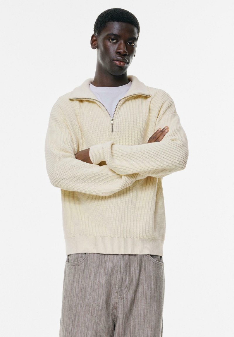 Cremefarbener, gerippter Reißverschluss-Pullover mit hohem Kragen und langen Ärmeln. Kombiniert mit hellgrauen gestreiften Hosen. Minimalistisches Design und weicher Stoff.