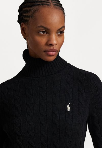 Svart kabelstickad polotröja med ribbad halsringning och en liten vit broderad logo på vänster sida. Mjuk, texturerad tyg.
