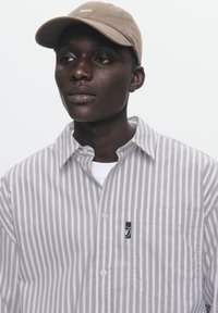 Striped button-up shirt in light gray and white, with a chest pocket and logo. Soft fabric; cap is beige with white embroidery.