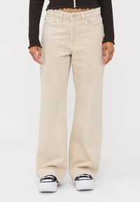 Beige vida jeans med hög midja, fem fickor, metallknappstängning och kontrasterande sömmar, kombinerade med svartvita sneakers.