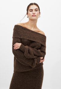 Maglione marrone a spalle scoperte realizzato in tessuto a maglia testurizzato, con maniche lunghe morbide e vestibilità rilassata. Il modello tiene le mani sui fianchi.