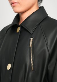 Giacca in pelle nera con un ampio colletto, bottone a scatto dorato e tasca con zip dotata di hardware metallici. Texture liscia e design aderente.