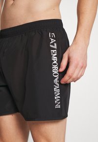 Black swim shorts with an elastic waist, featuring a white Emporio Armani logo printed vertically along the side. Smooth, lightweight material.