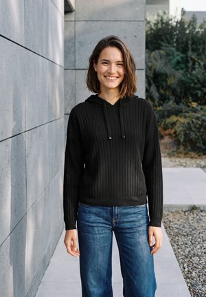 Une femme souriante se tient à l’extérieur, à côté d’un mur en béton. Elle porte un sweat à capuche noir côtelé et un jean bleu.