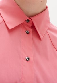 Primo piano di una persona che indossa una camicia rosa con colletto, con tre bottoni scuri visibili allacciati sul davanti.