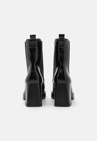 Botas de tobillo de charol negro con un grueso tacón cuadrado, lengüetas para facilitar el calce y una textura suave, que presentan una silueta elegante y sin patrones visibles.