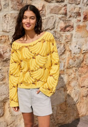 Kobieta ubrana w żółty sweter na drutach z wzorem bananów i jasnoszorty, stojąca przy strukturalnej kamiennej ścianie.