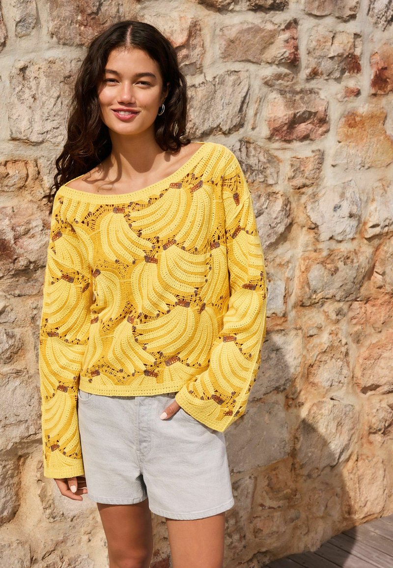 Femme portant un pull jaune en tricot avec des motifs de bananes et un short gris clair, debout contre un mur en pierre texturé.