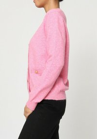 Cardigan rose fait d'un matériau doux, avec des manches longues, un col rond, deux poches avant et des détails en boutons dorés.