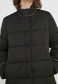 Czarna kurtka puchowa z wysokim kołnierzem, poziomym pikowaniem i złotym łańcuszkiem wokół dekoltu. Miękka, gładka faktura.