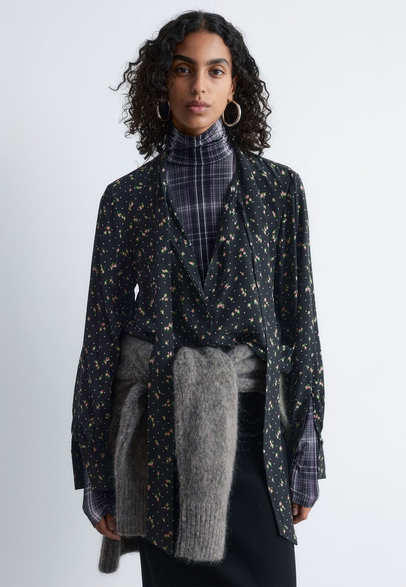 Floral black blouse with long sleeves, featuring a tie at the waist. Worn over a purple plaid turtleneck, paired with a gray wool sweater.