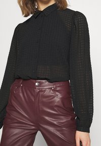 Camicia nera con bottoni, con un pattern testurizzato, maniche lunghe e colletto classico, abbinata a pantaloni in pelle sintetica bordeaux.