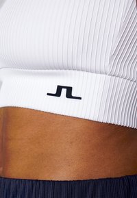 Bílý žebrovaný crop top s černým logem na pasu, zdůrazňující strukturovanou látku a přiléhavý střih, kombinovaný s tmavými plisovanými kalhotami.