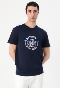Navy blue cotton T-shirt featuring a circular graphic with "Original Tommy Jeans New York" in white and red-striped letters, and year 1985.