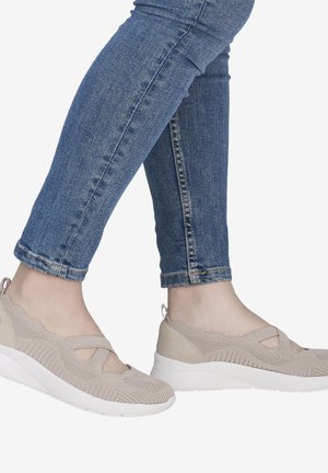Pieds chaussés de chaussures beige en maille sans lacets avec semelles blanches, associées à un jean skinny bleu sur un fond uni.
