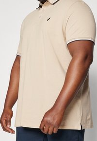 Beżowa koszulka polo z krótkimi rękawami, z czarnym logo na piersi, kontrastowym obszyciem przy kołnierzyku oraz rozcięciami po bokach na dole.