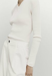 Haut rose clair à manches longues en côtes avec un col en V, associé à un pantalon blanc taille haute avec une fermeture par un seul bouton et des poches latérales.