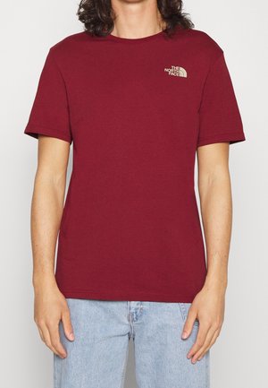Homme portant un t-shirt uni bordeaux à manches courtes avec le logo The North Face sur le haut gauche de la poitrine et un jean bleu clair.