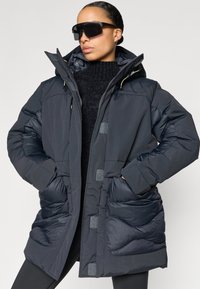 Mustast materjalist sulejope, millel on kapuuts, suured esimesed taskud kõverate õmblustega ja hallid detailid, valmistatud siledast materjalist.