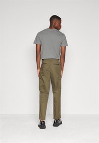 Graues Baumwoll-T-Shirt kombiniert mit olivgrünen Cargo-Hosen, die seitliche Taschen und einen strukturierten Stoff aufweisen. Schwarze Schuhe runden das Outfit ab.
