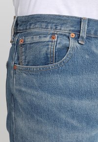 Jeans in denim azzurro chiaro con design a cinque tasche, rivetti in rame e cuciture visibili. Il tessuto ha una texture sbiadita e un taglio classico.