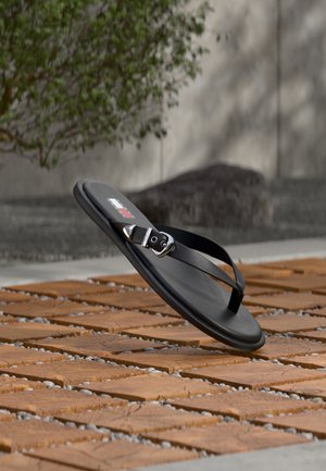 Zwarte string sandaal met zilveren gesp, hangend boven een houten tegelvloer buiten, met groene bladeren en een stenen muur op de achtergrond.