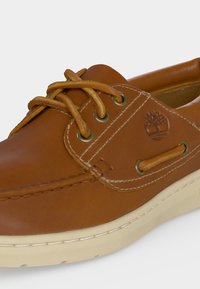 Chaussure bateau en cuir marron avec lacets couleur tabac, coutures blanches, œillets métalliques et semelle en caoutchouc blanche.