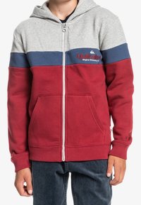 Persona che indossa una felpa con zip in blocchi di colore grigio, blu navy e rosso, con un logo rosso "Quiksilver" sul petto, abbinata a pantaloni scuri.
