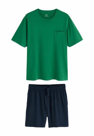 Mand med kort krøllet hår og skæg iført en grøn kortærmet T-shirt med en brystlomme, stående mod en ensfarvet grå væg.