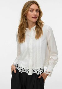 Femme aux longs cheveux blonds portant un chemisier blanc boutonné avec un bord en dentelle et un pantalon noir, les mains dans les poches, sur un fond blanc.