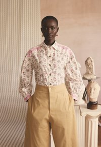 Femeie cu pielea închisă la culoare, cu păr scurt, purtând o bluză cu model floral și pantaloni bej cu talie înaltă, stând cu mâinile în buzunare lângă busturi sculptate.