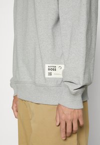 Felpa grigia a girocollo con polsini a costine, dotata di un'etichetta bianca sull'orlo con "BETTER BOSS" e un codice QR. Materiale in misto cotone.