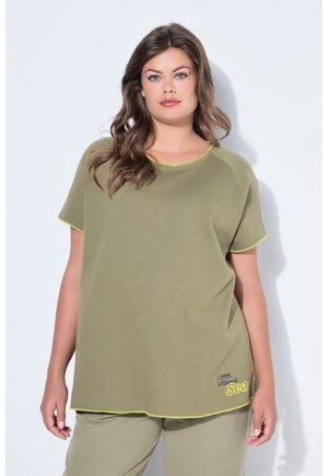 Donna che indossa un top a maniche corte e pantaloni verde oliva dalla vestibilità ampia, il top ha un bordo giallo e il testo "Ho bisogno di vitamina mare" vicino all'orlo.