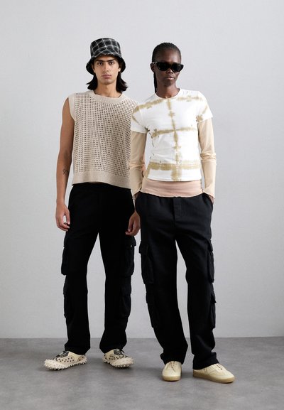 Top in maglia beige cropped a sinistra con pantaloni cargo neri; parte superiore crema tie-dye con maniche lunghe a destra con pantaloni neri; entrambi indossano calzature.