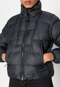 Giacca nera imbottita con alto colletto, chiusura con zip, polsini elasticizzati e tasche laterali, caratterizzata da una superficie liscia e lucida e un design trapuntato.