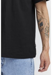 Black cotton t-shirt with short sleeves, featuring a side seam tag. Visible arm with detailed tattoos, including a playing card design.