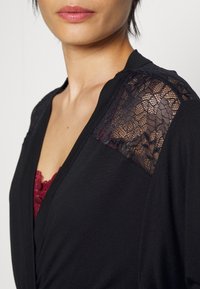 Vêtement noir avec un décolleté en V profond, épaules ornées de dentelle, et une texture qui contraste avec le tissu lisse. Détail en dentelle bourgogne visible.